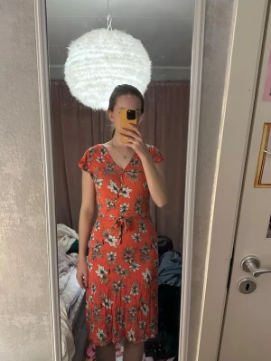 Röd blommig omlottklänning - Snygg röd omlottklänning med blommigt mönster och knyt i midjan. Klänningen har korta ärmar, v-ringning och faller mjukt ner till knäna. Perfekt för varma dagar och ger en fräsch sommarkänsla.