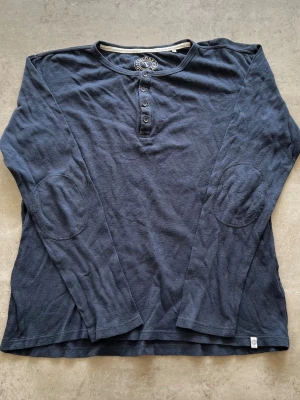 Mörkblå långärmad henleytröja - L - Mörkblå långärmad henleytröja från Ragwear med fyra knappar vid halsen och förstärkta armbågslappar. Tillverkad i mjukt, strukturerat bomullstyg som ger en avslappnad och skön känsla. Perfekt för lager på lager och enkel att matcha.