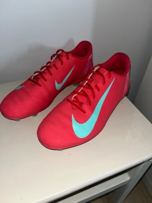 Röda Nike fotbollsskor med turkos swoosh - Snygga fotbollsskor från Nike i rött med turkosa detaljer och stor swoosh på sidan. Skorna har snörning, dobbar för bra grepp på planen och texten NIKE bak på hälen. Tillverkade i syntetmaterial för lätt känsla och bra bollkontroll. Använts en halv säsong bara.