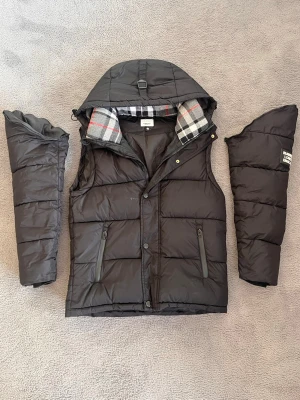 Svart dunjacka  - Svart dunjacka f med avtagbar huva och rutigt foder i grått, vitt och rött. Jackan har dragkedja, två fickor med dragkedja framtill och en modern, boxig passform. Den blir till väst ✅ Inga defekter på jackan ✅