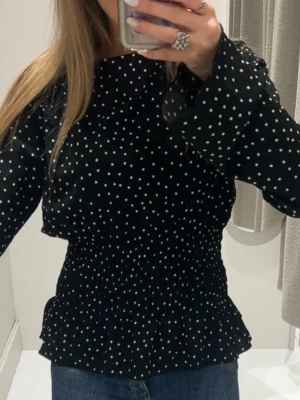 Svart prickig blus med smock - Trendig svart blus med vita små prickar och smockad midja som ger en snygg siluett. Blusen har långa ärmar med lätt utsvängda ärmslut och rund halsringning. Perfekt att styla med jeans eller kjol för en cool look.