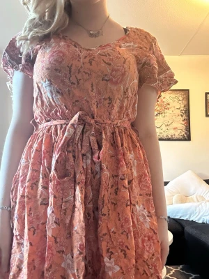 Blommig orange kortklänning med knyt - Söt kortklänning i orange med blommigt mönster och knytband i midjan. Klänningen har korta ärmar och v-ringad hals. Perfekt för varma dagar och ger en bohemisk vibe. Materialet känns lätt och luftigt, troligen bomull eller linne.