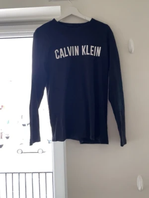 Svart långärmad topp Calvin Klein - Svart långärmad topp från Calvin Klein med vit logotyptext över bröstet. Klassisk rund halsringning och rak passform. Perfekt för en enkel och stilren look.