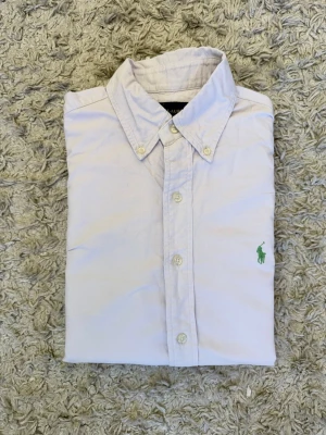 Vit skjorta från Ralph Lauren - Klassisk vit skjorta från Ralph Lauren med button-down krage och grönt broderat logotyp på bröstet. Skjortan har långa ärmar, knappar framtill och är tillverkad i mjuk bomull. Perfekt för en clean och stilren look.