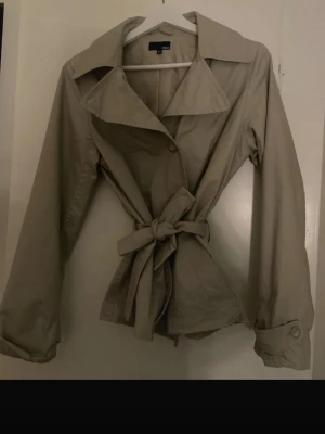 Beige kort trenchcoat med knytskärp - Stilren beigegrå kort trenchcoat från HM med klassisk krage, knytskärp i midjan och långa ärmar.  Storlek 40 men passar XS/S. Inga tecken på användning 