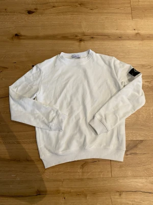 Vit sweatshirt från Stone Island - Säljer en stilren vit sweatshirt från Stone Island med klassisk rund hals och ribbade muddar. Tröjan har lång ärm och den ikoniska Stone Island-loggan på ärmen. Perfekt för dig som gillar enkel och clean stil.