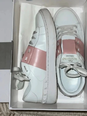 Valentino Garavani vita sneakers - De perfekta skorna för sommarn/våren köp nu när våren precis börjat och sommarn snart är här. Snygga vita sneakers från Valentino Garavani med bred rosa metallic detalj över mitten och vita snören. Skorna har en klassisk låg siluett och är tillverkade i slätt läder med subtila nitar på sulan. Perfekta för dig som gillar exklusiv och trendig stil.
