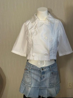 Vit kort jacka med trekvartsärm - Stilren vit kort jacka med dubbelknäppning och trekvartsärmar. Jackan har bred krage och dekorativa sömmar samt knappar både fram och bak. Perfekt för en trendig och fräsch look till våren eller sommaren.