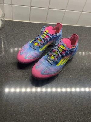Adidas f50 - Säljer ett par riktigt snygga Adidas fotbollsskor med en cool färgkombination av blått, rosa och neon-gult. Skorna har klassiska snörningar, låg profil och mönstrad sula för bra grepp på planen. Perfekta för dig som vill sticka ut på matchen!