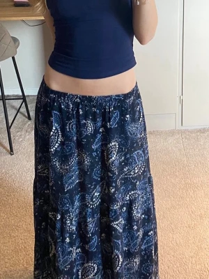 Mörkblå långkjol med paisleymönster - Säljer en mörkblå långkjol med vitt paisleyinspirerat mönster. Kjolen har en avslappnad passform med resår i midjan och är gjord i ett lätt och luftigt material. 93cm från midja och ner.