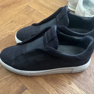 Axel arigato skor - Säljer ett par svarta slip-on sneakers från Axel Arigato med vit sula. Skorna är i mocka och har en stilren design utan snörning, med elastiskt band över vristen för enkel påtagning. Perfekta för en clean och modern look. Helt nya