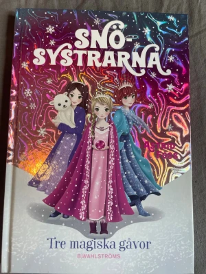 Snösystrarna - två böcker av Astrid Foss - Två färgglada böcker i serien Snösystrarna av Astrid Foss: 'Tre magiska gåvor' och 'Kristallrosen'. Omslagen har glittriga, vintriga illustrationer med tre systrar i magiska kläder och en vit isbjörn. Perfekt för dig som gillar äventyr och magi.