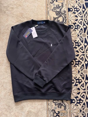 Svart sweatshirt från Polo Ralph Lauren - Klassisk svart sweatshirt från Polo Ralph Lauren med rund halsringning och diskret logobrodyr på bröstet. Tröjan har långa ärmar och ribbade muddar vid ärmslut och nederkant. Perfekt för en avslappnad och stilren look.