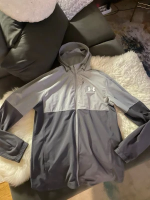 Grå och svart vindjacka Under Armour - Snygg vindjacka från Under Armour i tvåfärgad design, ljusgrå upptill och mörkgrå nertill. Jackan har huva, hel dragkedja framtill och Under Armour-logga på bröstet. Perfekt för blåsiga dagar och sportiga outfits.