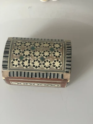 Mosaikmönstrad smyckesask i trä - Unik smyckesask i trä med handgjort mosaikmönster i svart, vitt och beige. Locket är välvt och insidan är klädd i röd sammet. Perfekt för att förvara ringar, örhängen eller andra småsaker. Asken har orientaliska detaljer och en klassisk känsla.