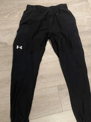 Svarta leggings från Under Armour - Säljer ett par svarta leggings från Under Armour med logga på vänster ben. De har en smal passform, elastisk midja och är tillverkade i ett stretchigt funktionsmaterial som passar perfekt till träning eller chill.