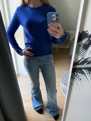 Kornblå stickad tröja med knappar - Snygg kornblå stickad tröja med rund hals och långa ärmar. Tröjan har dekorativa guldfärgade knappar vid ärmsluten som ger en extra detalj. Materialet ser tunt och mjukt ut, perfekt för lager på lager. Enkel och stilren design som passar till mycket.