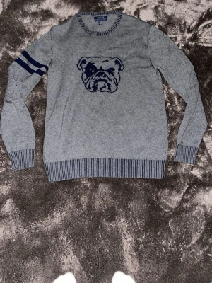 Polo Ralph Lauren tröja - Grå stickad tröja från Polo Ralph Lauren med en mörkblå bulldog på bröstet. Storlek xl i barnstorlek. Passar även folk som har storlek s i herrstorlek. Den har täcken på användning för det är lite knottrig på vissa ställen. 