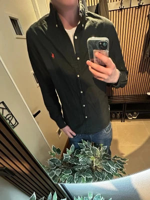 Mörkgrön skjorta från Ralph Lauren - Snygg mörkgrön skjorta från Ralph Lauren med klassisk button-down krage och den ikoniska röda logotypen broderad på bröstet. Skjortan har långa ärmar, vita knappar och är tillverkad i bomull. Perfekt till jeans för en clean look.