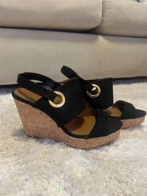 Svarta kilklackade sandaler med korksula - Säljer ett par svarta sandaler med breda band i ormmönstrad textil och stora guldfärgade ringdetaljer. Skorna har öppen tå och kilklack i korkmaterial som ger extra höjd och en cool look. Perfekta för sommaren och lätta att matcha med olika outfits.