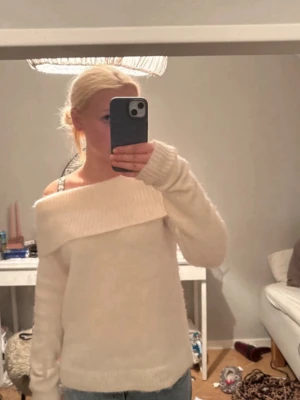 Off shoulder vit stickad tröja - Trendig off shoulder stickad tröja i mjuk vit färg. Tröjan har bred ribbad kant upptill som ger en snygg axelbar look och långa ärmar. Perfekt för dig som vill ha en stilren och modern vibe. Passar till både jeans och kjol.