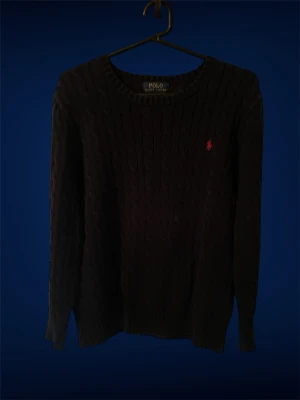 Mörkblå kabelstickad tröja Polo Ralph Lauren - Kabelstickad mörkblå tröja från Polo Ralph Lauren med rund halsringning och lång ärm. Tröjan har det klassiska röda Polo-logot på bröstet och ribbade muddar vid ärmslut och nederkant. Perfekt för dig som gillar stilren och tidlös design.