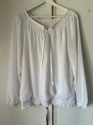 Vit blus med smock och knytband - Luftig vit blus med smockdetaljer vid halsringning och nederkant samt knytband framtill. Blusen har långa ärmar med resår vid ärmslut och är tillverkad i ett tunt, lätt material som ger en avslappnad look.