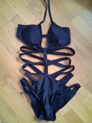 Svart cut-out baddräkt med band - Säljer en svart baddräkt med flera cut-out detaljer och smala band över magen och sidorna. Halterneck-modell med vadderade kupor och trendig design som sticker ut på stranden eller vid poolen. Perfekt för dig som vill ha en unik och snygg baddräkt.