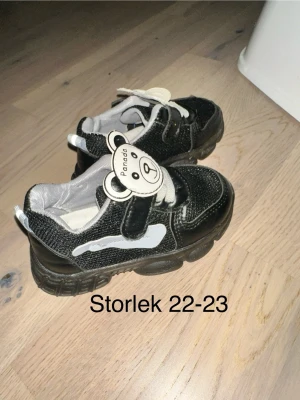 Svarta sneakers med panda-detalj - Säljer ett par svarta sneakers med vit panda-detalj på kardborrebandet och vita detaljer på sidan. Skorna har en chunky sula och är tillverkade i textil och syntetmaterial. Perfekta för dig som gillar gulliga detaljer och bekväma skor.