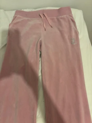 Rosa velourbyxor Juicy Couture - Supermjuka rosa velourbyxor från Juicy Couture med bred resårmidja och dragsko. Byxorna har strassdetaljer med JC-logga framtill och Juicy Couture-text bak. Perfekta för chill dagar och en ikonisk Y2K-look.