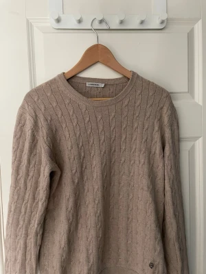 Beige kabelstickad tröja J.Lindeberg - Kabelstickad beige tröja från J.Lindeberg med rund halsringning och diskret logga vid nederkanten. Tröjan har långa ärmar och ribbade muddar. Perfekt för lager på lager och enkel att matcha med jeans eller kjol. 100% bomull. Storlek M.