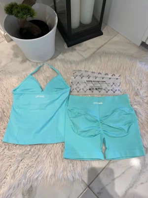 ICE DFYNE - Säljer ett turkost träningsset från DFYNE med ett halterneck-linne och matchande shorts. Setet har vita logotyper och är gjort i ett stretchigt, mjukt material som passar perfekt för gym eller yoga. Snygg passform och sportig look. Modell: impact 4.5 i färgen ICE
