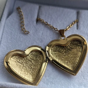Medaljong Guldfärgat hjärthalsband med gravyr - Säljer ett guldfärgat hjärthalsband med vackra ingraverade blommönster. Halsbandet har en kedja i rostfritt stål med justerbar längd och hjärtberlocken går att öppna för att lägga in en liten bild eller minne. Perfekt accessoar för dig som gillar romantiska detaljer. Kedja 45 cm och hänget ca 3 cm. 