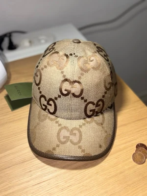 Beige keps från Gucci med GG-mönster - Snygg beige keps från Gucci med det klassiska GG-monogrammet i brunt över hela kepsen. Kepsen har böjd skärm och är tillverkad i ett slitstarkt tygmaterial. Perfekt för dig som vill ha en exklusiv och trendig accessoar. Skriv gärna om ni har frågor svarar direkt
