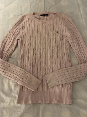 Ljusrosa kabelstickad tröja Gant - Supersnygg ljusrosa kabelstickad tröja från Gant med rund halsringning och diskret logga på bröstet. Tröjan har långa ärmar och ribbade muddar vid ärmslut och nederkant. Perfekt för dig som gillar klassisk och stilren look.