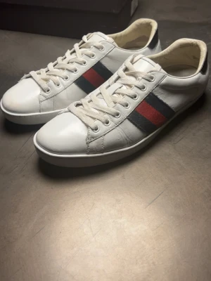 Gucci skor - Snygga vita sneakers från Gucci i läder med klassiska röd och blå ränder på sidorna. Skorna har vita snören och låg profil, perfekt för en stilren och trendig look. Ikonisk design som passar till många outfits.