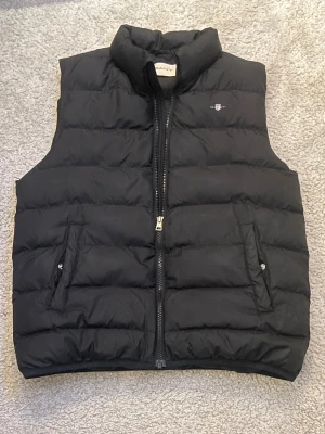Svart dunväst från GANT  - Väldigt fin dunväst från gant, bra skick förutom ett litet hål i högra armvecket (syns på bild 4) inget dun läcker ut och syns ej vid användning. passar bra om man är runt 160cm. Hör av er vid funderingar!