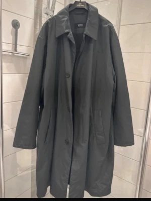 Svart rock från Hugo Boss - Stilren svart rock från Hugo Boss med klassisk krage och knäppning framtill. Rocken har två sidofickor och är tillverkad i ett slätt material med diskret glans. Perfekt för dig som vill ha en tidlös och snygg look.
