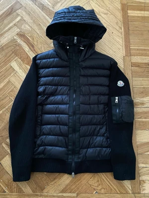 Svart pufferjacka från Moncler - Snygg svart pufferjacka från Moncler med quiltad front och huva. Jackan har stickade ärmar och rygg, samt en praktisk ficka med dragkedja på ena ärmen. 🚨🚨🚨Dom kommer lite små i storlek så de som brukar ha på sig storlek M skulle passa perfekt. va inte blyg o fråga ❤️