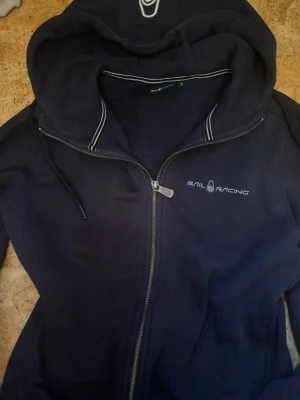Marinblå hoodie från Sail Racing - Snygg marinblå hoodie från Sail Racing med dragkedja framtill och vit logga på bröstet. Huva med dragsko och vita detaljer längs kanten. Mjuk insida och klassisk passform, perfekt för chill dagar eller när du vill ha en sportig look.