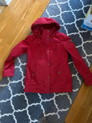 Röd regnjacka med huva - Snygg röd regnjacka med huva och dragsko. Jackan har tryckknappar framtill, två fickor med lock och detaljer i metall. Insidan av huvan har ett mönstrat tyg. Perfekt för regniga dagar och enkel att matcha med olika outfits.