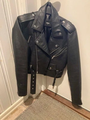 Svart skinnjacka från Bik Bok XS - Säljer en svart skinnjacka från Bik Bok i storlek XS. Klassisk bikerstil med sned dragkedja, axelklaffar, bälte i midjan och flera fickor med dragkedja. Med silverdetaljer.