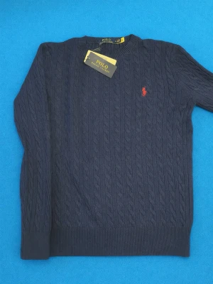  tröja Polo Ralph Lauren - Mörkblå kabelstickad tröja från Polo Ralph Lauren med rund halsringning och den klassiska röda logotypen broderad på bröstet. Tröjan har långa ärmar och ribbade muddar vid ärmslut och nederkant. Perfekt för en stilren och tidlös look.