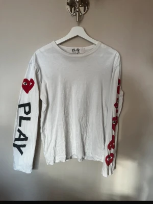Vit långärmad Comme des Garçons - Vit långärmad topp från Comme des Garçons PLAY med svart text och röda hjärtan på ärmarna. Ikoniskt hjärtmotiv med ögon på ena ärmen och PLAY-tryck. Tillverkad i mjuk bomull med relaxed fit och rund hals.