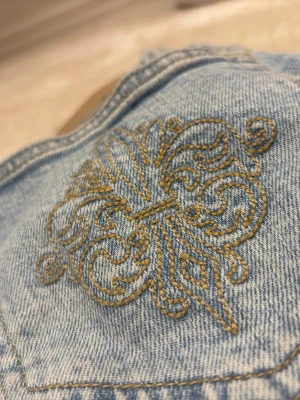 Ljusblå bootcut jeans med brodyr - Snygga ljusblå bootcut jeans med dekorativt broderi i gult på bakfickan. Jeansen har klassisk femficksdesign och är tillverkade i denim med lätt tvättad look. Perfekta för en avslappnad och trendig stil.