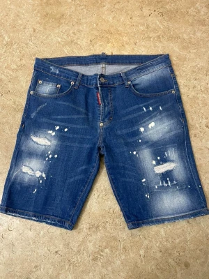 Blå jeansshorts från Dsquared2 - Säljer ett par blå jeansshorts från Dsquared2 med slitna detaljer och vita färgstänk. Klassisk femficksmodell med knappgylf och bälteshällor. Shortsen har en avslappnad passform och är tillverkade i bomull, perfekta för sommaren.