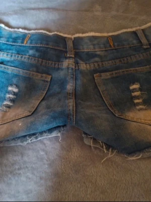 Blå slitna jeansshorts med fransar - Säljer ett par blå lågmidjade jeansshorts med slitna detaljer och fransig kant. De är lite små i storleken så de är egentligen M men skulle säga att de sitter bättre på S. Midjemåttet är 37cm rakt över. Shortsen har klassisk femficksdesign och knappar framtill. Perfekta för sommaren och ger en cool, avslappnad vibe.