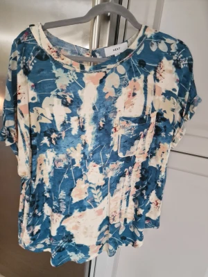Blommig blå och vit blus från Next - Säljer en luftig blus från Next med blått och vitt blommönster, rund halsringning och korta ärmar. Toppen har en liten bröstficka och knappdetalj i nacken. Perfekt för dig som gillar en avslappnad men ändå snygg stil.