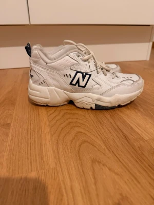 Vita New Balance 608 sneakers - Chunky vita New Balance 608 sneakers med blå detaljer och klassisk logga på sidan och hälen. Skorna har en robust sula och är tillverkade i läder med meshpaneler för extra komfort. Perfekta för dig som gillar retrostil och streetwear.