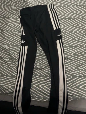 Svarta Adidas leggings med vita ränder - Säljer ett par svarta Adidas leggings med klassiska vita ränder längs benen och Adidas-logga på låret. Tight passform och elastisk midja, perfekta för en sportig och avslappnad stil.